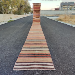 May include: A long, narrow runner rug with a striped pattern in shades of beige, brown, and orange. The rug is displayed outdoors on a paved road, showcasing its length and design. The rug's fringe is visible at the end.
