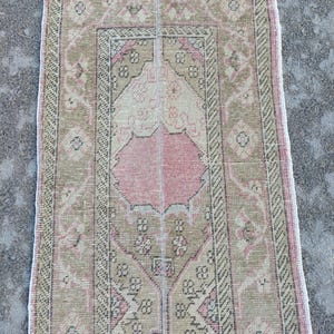 May include: A rectangular, vintage rug with a central pink medallion and a border of repeating geometric patterns. The rug features a colour palette of muted pink, beige, and olive green, with a distressed appearance.