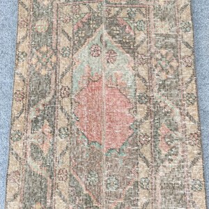 Peut inclure: Un tapis rectangulaire vieilli avec un motif de médaillon central. Le tapis présente une palette de couleurs de verts sourds, de bruns et de rouges délavés. Les bords ont un détail effiloché, et l'esthétique générale est vintage.