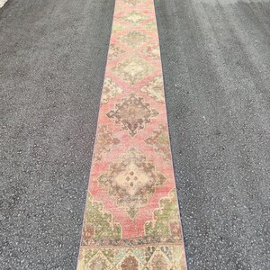 May include: A long, rectangular runner rug with a faded, vintage appearance. The rug features a central field of faded red with intricate geometric patterns in beige, brown, and green. The rug is placed on a dark grey surface.