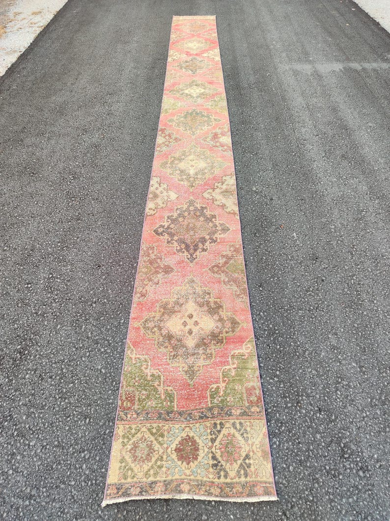 May include: A long, rectangular runner rug with a faded, vintage appearance. The rug features a central field of faded red with intricate geometric patterns in beige, brown, and green. The rug is placed on a dark grey surface.