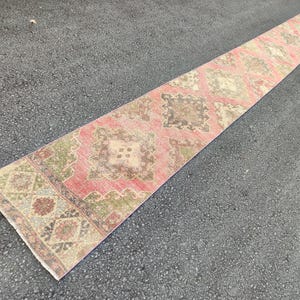 May include: A long, rectangular runner rug with a faded, vintage appearance. The rug features a repeating pattern of geometric shapes in shades of pink, brown, green, and beige. The rug is placed on a dark grey surface.