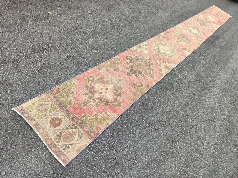 May include: A long, rectangular runner rug with a faded, vintage appearance. The rug features a repeating pattern of geometric shapes in shades of pink, brown, green, and beige. The rug is placed on a dark grey surface.