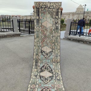 May include: A long, rectangular rug with an intricate pattern. The rug features a central design with diamond shapes in black, cream, and blue, surrounded by floral and geometric motifs in muted colors. The rug is held upright, showcasing its length and design.
