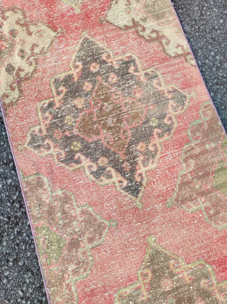 May include: A distressed, rectangular rug with a faded, vintage appearance. The rug features a central medallion design in shades of black, brown, and pink, surrounded by a pink and beige background. The rug's edges are a lighter pink.