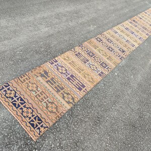 May include: A long, rectangular runner rug with a geometric pattern. The rug features a repeating design in shades of beige, blue, purple, and orange. The rug is placed on a gray surface, showcasing its length and intricate details.