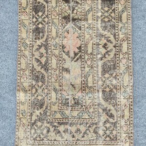 Könnte beinhalten: Ein rechteckiger Vintage-Teppich mit einem Used-Look. Er hat ein zentrales Blumendesign in Rosa und Beige, umgeben von geometrischen Mustern in Braun, Grün und Creme. Die Kanten sind leicht ausgefranst, was ihm einen antiken Charme verleiht. Geeignet für einen Flur.