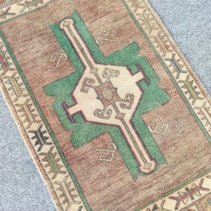 May include: A rectangular, vintage rug with a central design in green and cream, set against a brown background. The rug features a detailed border with geometric patterns and a faded, antique appearance.