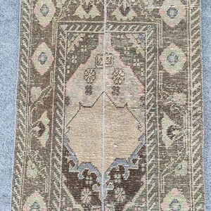 May include: A rectangular, vintage rug with a central medallion design. The rug features a palette of brown, beige, and muted green, with accents of pink and blue. The border has a geometric pattern, and the rug appears to be made of woven material.