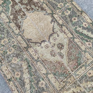 Peut inclure: Un tapis rectangulaire vieilli et délavé, d'aspect vintage. Le tapis présente un médaillon central beige entouré d'une bordure de motifs floraux verts, bruns et beiges. Le design du tapis a une esthétique antique et usée.