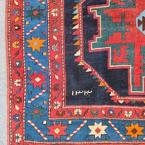 Puede incluir: Una alfombra tejida que presenta un campo central azul profundo con un gran medallón geométrico rojo y verde. El borde muestra un patrón vibrante de secciones rojas y azules adornadas con motivos de estrellas multicolores y flores.