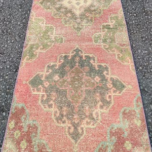 May include: A rectangular, distressed rug with a pink backdrop and detailed patterns in green, grey, and burgundy. The rug's design includes a central medallion and corner details, giving it a vintage appearance. The rug is suitable for a hallway.
