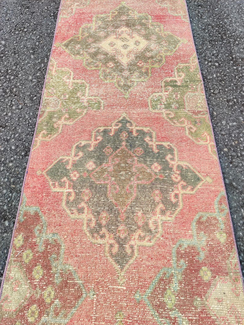 May include: A rectangular, distressed rug with a pink backdrop and detailed patterns in green, grey, and burgundy. The rug's design includes a central medallion and corner details, giving it a vintage appearance. The rug is suitable for a hallway.