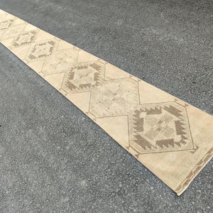 May include: A long, rectangular beige runner rug with geometric patterns in shades of brown. The rug features repeating diamond and hexagonal shapes, creating a symmetrical design. The rug is placed on a grey surface.