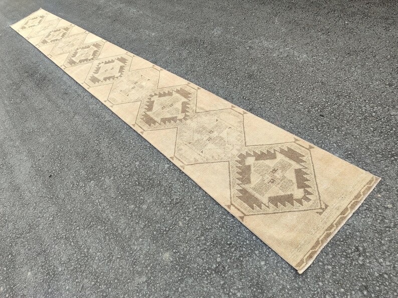 May include: A long, rectangular beige runner rug with geometric patterns in shades of brown. The rug features repeating diamond and hexagonal shapes, creating a symmetrical design. The rug is placed on a grey surface.