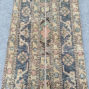May include: A vintage runner rug with a distressed, faded appearance. The rug features a repeating pattern of floral and geometric motifs in shades of navy blue, beige, and muted pink. The rug's design is symmetrical, with vertical stripes.