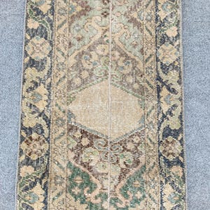 Op de afbeelding: Een rechthoekig vloerkleed in vintage-stijl met een centraal ruitpatroon. Het vloerkleed heeft een beige achtergrond met ingewikkelde bloemen- en geometrische ontwerpen in tinten groen, bruin en marineblauw. De randen zijn afgezet met een donkerder marineblauw.