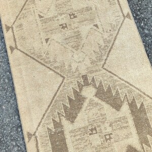 May include: A beige and brown patterned rug with geometric designs. The rug features a central diamond shape with repeating patterns and a border of triangles. The colour palette is neutral, with shades of beige and brown.