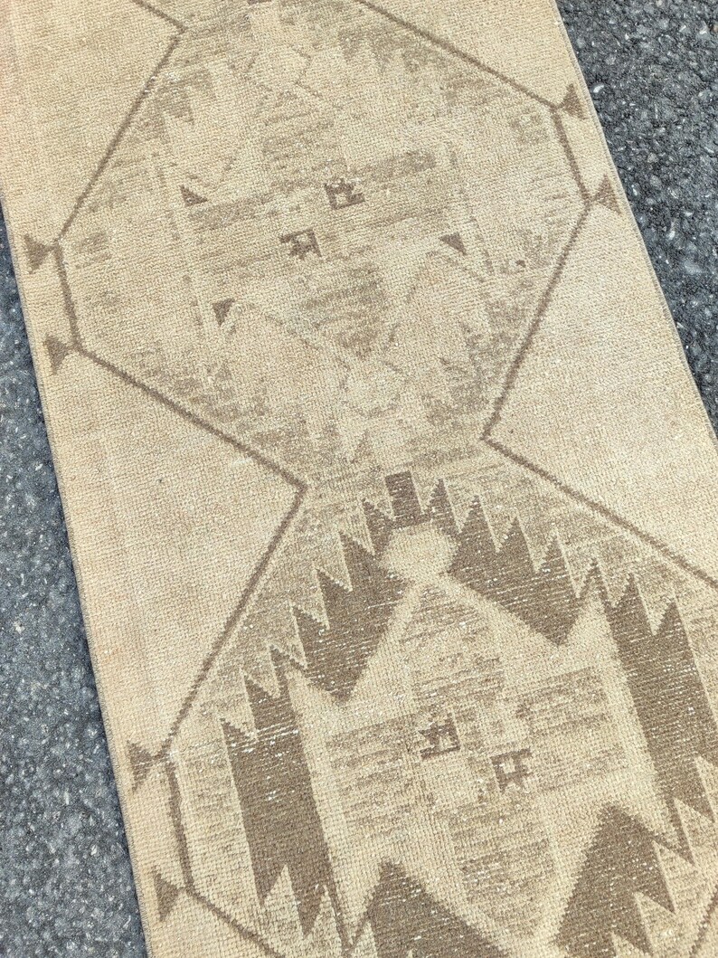 May include: A beige and brown patterned rug with geometric designs. The rug features a central diamond shape with repeating patterns and a border of triangles. The colour palette is neutral, with shades of beige and brown.
