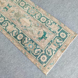 May include: A rectangular rug with a detailed, geometric pattern. The rug features a central beige field with a border of teal and tan. The design includes floral and geometric motifs. The rug is placed on a grey surface.