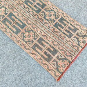 Könnte beinhalten: Ein rechteckiger Teppich mit einem geometrischen Muster in den Farbtönen Türkis, Pfirsich und Braun. Der Teppich weist ein sich wiederholendes Design aus stilisierten Formen und Linien auf, mit einem Rand in einem dunkleren Rotton. Der Teppich liegt auf einer grauen Oberfläche.