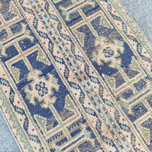 Könnte beinhalten: Nahaufnahme eines Abschnitts eines Vintage-Teppichs mit geometrischem Muster. Der Teppich weist eine Kombination aus blauen, beigen und grünen Farben auf. Das Design umfasst sich wiederholende Formen und florale Motive, wodurch ein strukturiertes Aussehen entsteht.