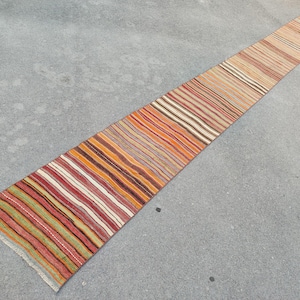 May include: A long, narrow runner rug with a striped pattern in shades of red, orange, brown, and cream. The rug has a fringed edge and is likely made of woven fabric. The design features a mix of vertical stripes.