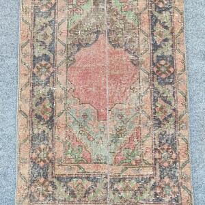Op de afbeelding: Een rechthoekig, versleten tapijt met een centraal ruitpatroon in tinten koraal en perzik. Het tapijt heeft een rand met bloemen en geometrische ontwerpen in groen, zwart en bruin. Het tapijt heeft een vintage, versleten uitstraling.