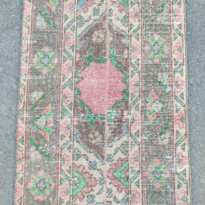 Könnte beinhalten: Ein rechteckiger Vintage-Teppich mit einem Used-Look. Der Teppich hat eine zentrale rosa Diamantenform, umgeben von braunen, beigen und grünen geometrischen Mustern. Das Design umfasst Blumen- und Diamantmotive.
