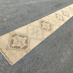 May include: A long, rectangular beige runner rug with a repeating geometric pattern in shades of brown. The rug is placed on a grey surface, showcasing its length and design. The pattern features diamond and square shapes.