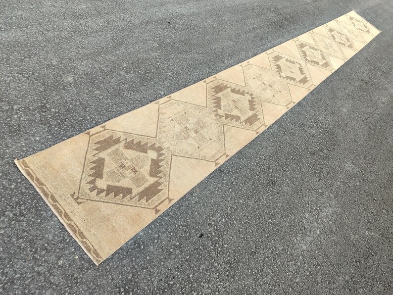 May include: A long, rectangular beige runner rug with a repeating geometric pattern in shades of brown. The rug is placed on a grey surface, showcasing its length and design. The pattern features diamond and square shapes.