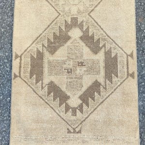 May include: A rectangular rug in beige tones, featuring a central geometric design in brown shades. The pattern includes diamond shapes and angular motifs. The rug has a distressed, vintage look, with a fringed edge along the bottom.