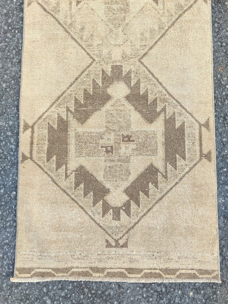 May include: A rectangular rug in beige tones, featuring a central geometric design in brown shades. The pattern includes diamond shapes and angular motifs. The rug has a distressed, vintage look, with a fringed edge along the bottom.