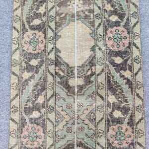 Könnte beinhalten: Ein Teppich im antiken Stil mit einem Used-Look. Der Teppich weist ein zentrales Rautenmuster und florale Motive in Beige-, Braun-, Grün- und Rosatönen auf. Das Design ist symmetrisch, mit vertikalen Streifen und einer verblichenen Vintage-Ästhetik.