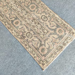 Op de afbeelding: Een rechthoekig vloerkleed in vintage stijl met een vervaagde, versleten uitstraling. Het vloerkleed heeft een centraal ontwerp met bloemen- en geometrische patronen in gedempte tinten grijs, beige en roze. De rand is een donkerdere tint met ingewikkelde details.