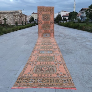 May include: A long, rectangular, vintage rug with a peach background and intricate geometric patterns in shades of green, blue, and brown. The rug is displayed outdoors on a stone walkway, showcasing its length and design.