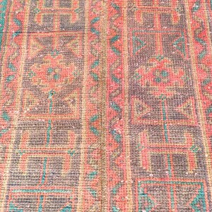 Könnte beinhalten: Nahaufnahme eines rechteckigen, gemusterten Teppichs mit einem sich wiederholenden geometrischen Design. Der Teppich hat eine Farbpalette aus Braun, Koralle und Türkis. Die Textur des Teppichs scheint gewebt zu sein, mit sichtbaren Fäden und einem leicht abgenutzten Aussehen.