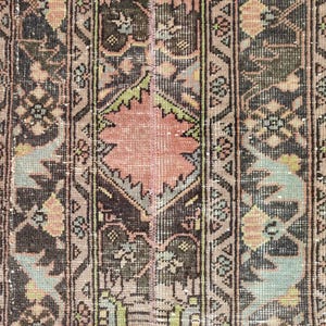 May include: Close-up of a distressed, rectangular rug with a central floral design in shades of peach and green. The rug features a repeating pattern of geometric shapes and floral motifs in brown, black, and muted green tones. The rug has a vintage, worn appearance.