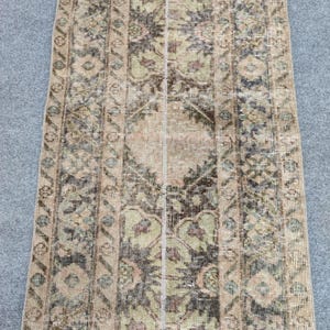 May include: A long, rectangular runner rug featuring a faded, distressed pattern in shades of beige, sage green, and dark brown. The design includes a central medallion motif flanked by intricate geometric borders with repeating floral elements.