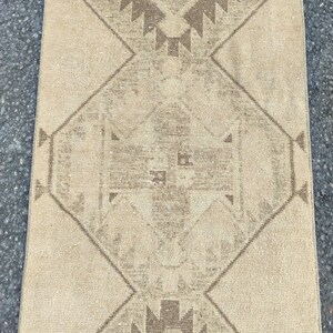 May include: A rectangular, beige rug with a geometric pattern. The design features diamond shapes and zigzags in shades of brown. The rug has a distressed, vintage appearance, suitable for home decor.