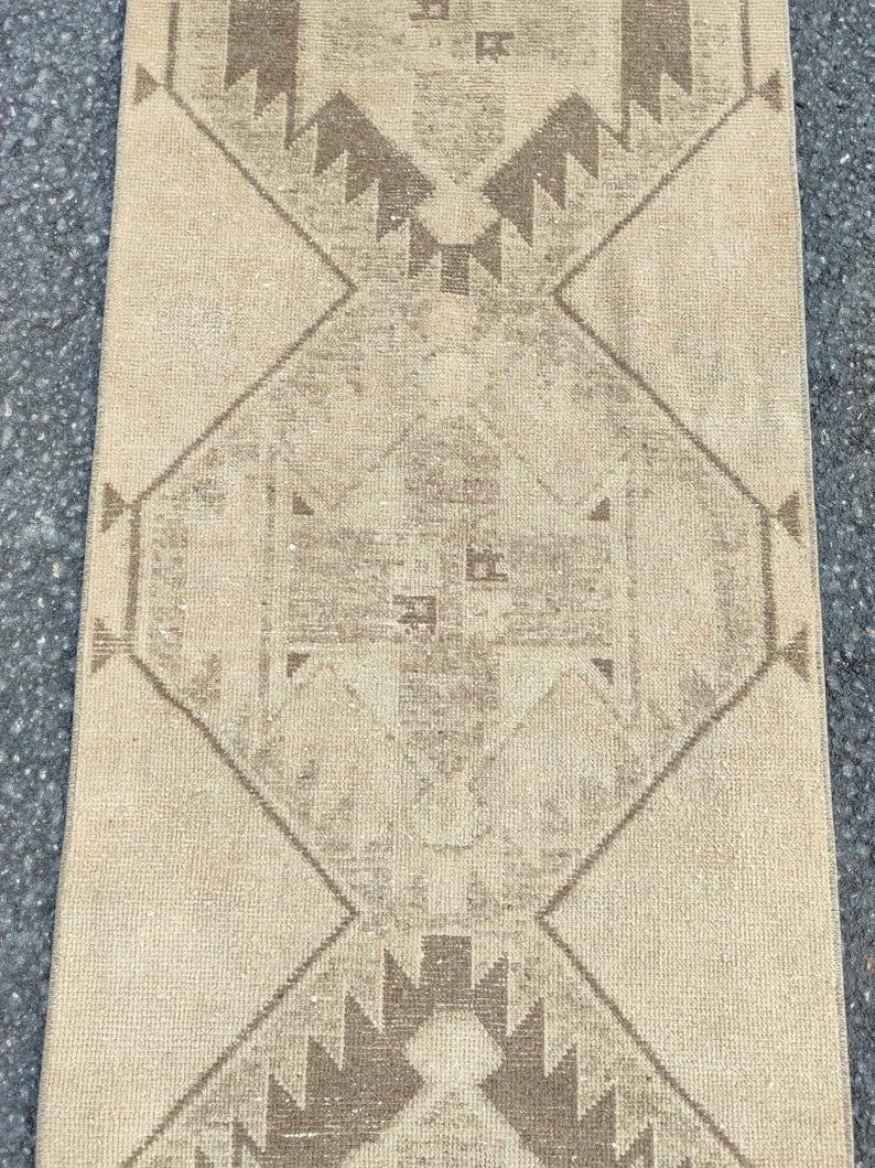 May include: A rectangular, beige rug with a geometric pattern. The design features diamond shapes and zigzags in shades of brown. The rug has a distressed, vintage appearance, suitable for home decor.