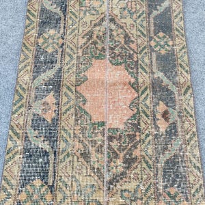 Könnte beinhalten: Ein rechteckiger, abgenutzter Teppich mit Patchwork-Design. Der Teppich hat einen zentralen pfirsichfarbenen Bereich, der von geometrischen Mustern in Schwarz-, Grün- und Beigetönen umgeben ist. Das gealterte Aussehen des Teppichs deutet darauf hin, dass er Vintage ist oder eine abgenutzte Ästhetik hat.