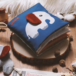 May include: A blue fabric baby book featuring a white elephant design with a red ear. The book is stacked on a wooden slice, with other baby toys and cotton balls in the background. The book has the word "oooo" printed on the top left corner.