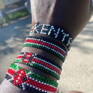 Könnte beinhalten: Mehrere Armbänder aus schwarzen, roten, weißen und grünen Perlen. Ein Armband schreibt "KENYA" mit weißen Perlen. Die Armbänder zeigen Muster und Farben, die an die kenianische Flagge erinnern.