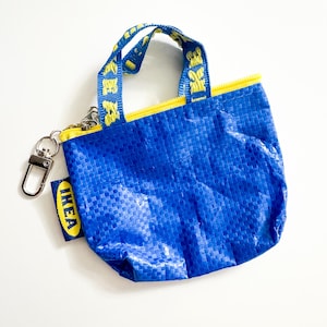 Può includere: Piccola borsa tote blu con finiture e manici gialli. La borsa ha una trama intrecciata e una chiusura a cerniera. I manici sono blu con testo giallo. Un moschettone metallico e un'etichetta "IKEA" sono attaccati.