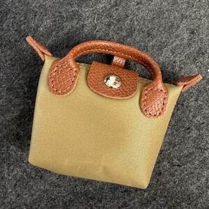 May include: A small, tan handbag with brown leather accents. The bag features a top handle and a flap closure with a silver-toned button. The bag's design is simple and elegant, suitable for various occasions.