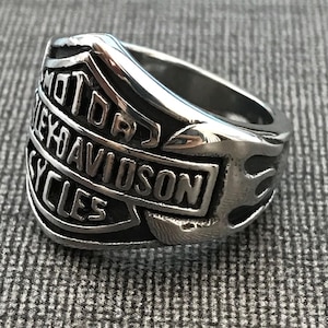 Op de afbeelding: Zilverkleurige ring met de woorden "MOTOR HARLEY-DAVIDSON CYCLES" in reliëf. De ring heeft een vlammenontwerp aan de zijkant. De achtergrond is een grijze, getextureerde stof.