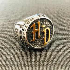 Könnte beinhalten: Ein silberfarbener Ring mit einem kreisförmigen Gesicht, das die goldfarbenen Buchstaben "HD" zeigt. Der Ring trägt die Inschrift "MOTORCYCLES" um das Band. Der Ring liegt auf einer grauen, strukturierten Oberfläche.