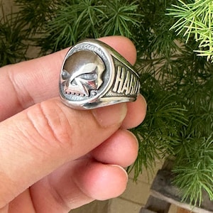 Peut inclure: Bague en argent ornée d'un crâne et du mot "HARD" gravé sur le côté. La bague présente une esthétique vintage détaillée. L'arrière-plan est composé de feuillage vert.