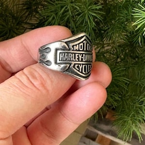 Può includere: Anello color argento con il logo Harley-Davidson. L'anello presenta un'emblema rettangolare con il nome del marchio e la parola "CYCLES" sottostante. Le fiamme sono incise sui lati dell'anello. L'anello è tenuto contro uno sfondo di fogliame verde.